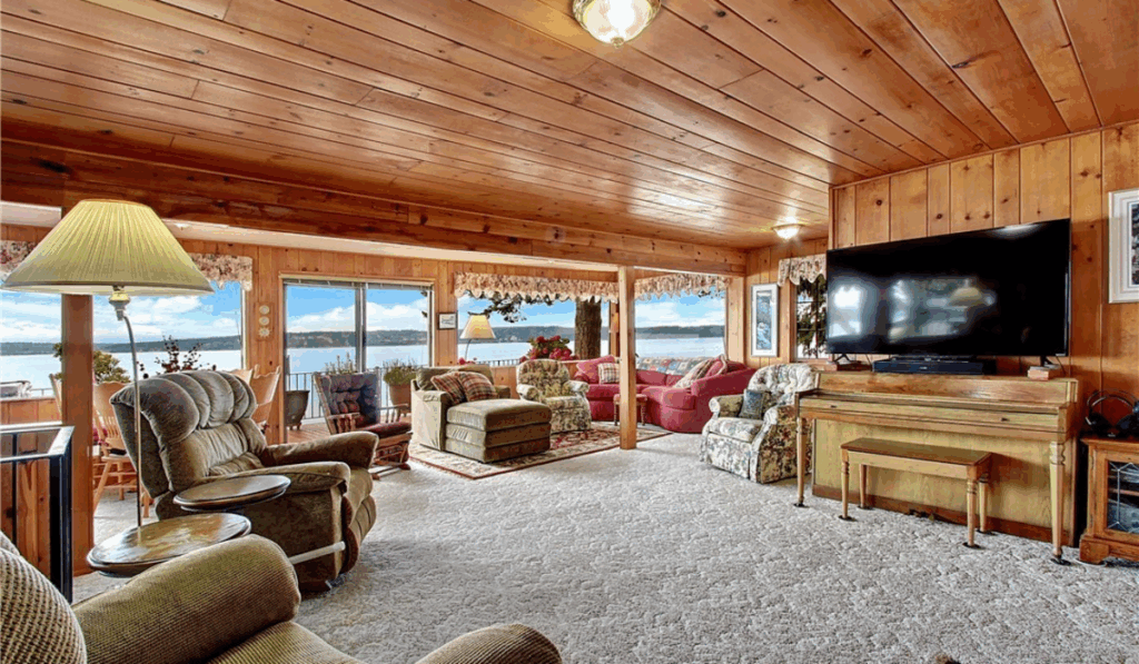 Charming waterfront living room with wood-paneled walls and ceiling, cozy carpet, ample seating, large TV, and stunning panoramic Puget Sound views—perfect for relaxing or entertaining